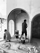 Two individuals are working in a construction or renovation setting. One person is crouched down holding a tool, while the other is standing and focused on a task. The setting is indoors, and unfinished walls with arches are visible. There are various construction materials and equipment scattered on the floor, including paint buckets, plastic sheets, and wooden planks.