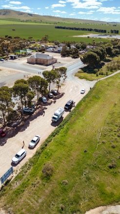 A rural landscape with a small intersection, several parked cars on a dirt road, and a few people walking nearby. A house and some auxiliary buildings are visible, surrounded by trees. In the background, vast green fields stretch out with wind turbines on the hills.