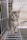 A small, fluffy kitten with blue eyes sits inside a wire cage. The kitten has a soft gray and white striped fur coat and is sitting on a cloth or blanket. Behind the kitten, a bowl of dry cat food is visible inside the cage.