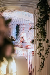 A beautifully decorated indoor space features a three-tiered cake surrounded by floral arrangements and adorned with lights. The setting includes intricate lattice work and is adorned with greenery, creating an elegant and festive atmosphere.