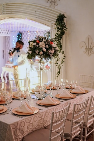 A beautifully set dining table with white chairs, covered with a patterned tablecloth and adorned with elegant glassware and neatly folded napkins on gold plates. A floral centerpiece with white, green, and red elements stands prominently on the table. The background features decorative arches, greenery, and warm lighting. A person dressed in light-colored formal attire is speaking into a microphone nearby.
