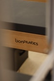 A close-up view of a Pilates reformer bearing the brand name 'bonPILATES'. The equipment is made of light-colored wood with a dark padded surface.