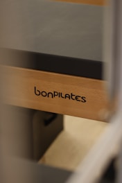 A close-up view of a Pilates reformer bearing the brand name 'bonPILATES'. The equipment is made of light-colored wood with a dark padded surface.