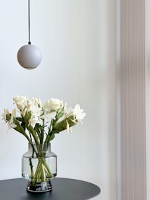A minimalist room corner features a round, grey pendant light hanging above a bouquet of white flowers in a transparent glass vase, which sits on a round black table. The background consists of a soft, neutral-colored wall with subtle vertical textures.