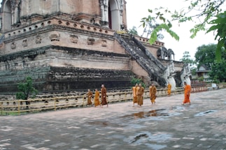 The image depicts several monks dressed in saffron robes walking around an ancient, large, and weathered brick temple structure. The temple features an elaborate staircase guarded by large, sculpted figures resembling mythical creatures. There's lush greenery in the background, and the ground appears damp, suggesting recent rain.