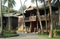 The photo shows a two-story wooden building with traditional-style railings and decorative elements. The structure is surrounded by lush tropical vegetation, including several palm trees. The foreground features a paved path leading to the entrance, with a small garden area featuring various plants and shrubs. Natural sunlight gives the scene a warm, serene atmosphere.