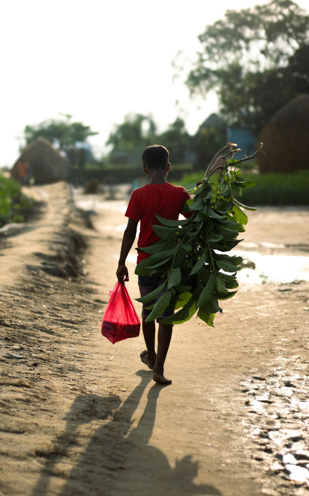 a boy walking down a dirt road carrying a red bag