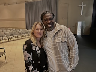A man and a woman are standing close together, smiling, in an indoor space that resembles a church or auditorium. The room has rows of empty chairs, a large curtain, and a cross mounted on the wall.