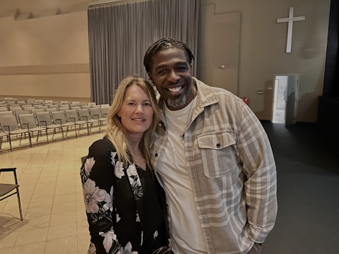 A man and a woman are standing close together, smiling, in an indoor space that resembles a church or auditorium. The room has rows of empty chairs, a large curtain, and a cross mounted on the wall.