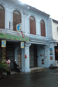 A vintage-style two-story building with light blue walls and wooden shutters. The establishment is marked by signage for a foot reflexology business. There are decorative details and columns on the facade. A person is standing near the entrance, next to a motorbike and several potted plants.