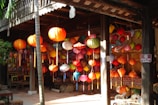 A collection of colorful lanterns hangs in a wooden market stall, illuminated by sunlight. The scene is vibrant, with lanterns in various shapes and sizes, displayed in a lively arrangement. In the background, there are wooden benches and a lush green bamboo plant adding a natural touch to the setting.