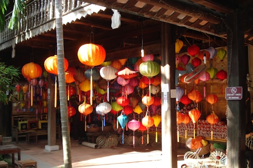 A collection of colorful lanterns hangs in a wooden market stall, illuminated by sunlight. The scene is vibrant, with lanterns in various shapes and sizes, displayed in a lively arrangement. In the background, there are wooden benches and a lush green bamboo plant adding a natural touch to the setting.