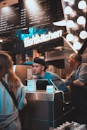 A busy kitchen scene with two workers behind a counter. One worker is engaging with a customer, while another worker is standing beside him. There is a prominent 'fish kitchen' sign above them, and a menu board showing various dish options and prices. The counter has a tip jar and a cash register.
