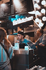 A busy kitchen scene with two workers behind a counter. One worker is engaging with a customer, while another worker is standing beside him. There is a prominent 'fish kitchen' sign above them, and a menu board showing various dish options and prices. The counter has a tip jar and a cash register.