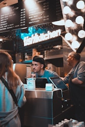 A busy kitchen scene with two workers behind a counter. One worker is engaging with a customer, while another worker is standing beside him. There is a prominent 'fish kitchen' sign above them, and a menu board showing various dish options and prices. The counter has a tip jar and a cash register.