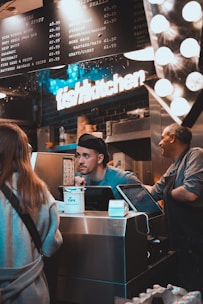 A busy kitchen scene with two workers behind a counter. One worker is engaging with a customer, while another worker is standing beside him. There is a prominent 'fish kitchen' sign above them, and a menu board showing various dish options and prices. The counter has a tip jar and a cash register.