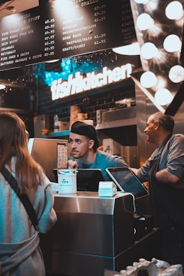 A busy kitchen scene with two workers behind a counter. One worker is engaging with a customer, while another worker is standing beside him. There is a prominent 'fish kitchen' sign above them, and a menu board showing various dish options and prices. The counter has a tip jar and a cash register.