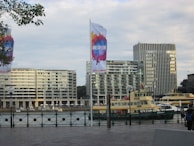 An urban waterfront scene featuring modern high-rise buildings and a harbor with ferries docked nearby. Flags with colorful designs and the word 'INSPIRE' prominently hang from large poles. Trees and foliage are visible in the foreground, adding a touch of nature to the cityscape.