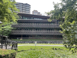 A modern multi-story building surrounded by greenery. The structure features horizontal lines and large glass windows, blending with the natural environment. There are trees and bushes in the foreground and background, providing a serene and natural setting.
