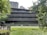 A modern multi-story building surrounded by greenery. The structure features horizontal lines and large glass windows, blending with the natural environment. There are trees and bushes in the foreground and background, providing a serene and natural setting.