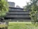 A modern multi-story building surrounded by greenery. The structure features horizontal lines and large glass windows, blending with the natural environment. There are trees and bushes in the foreground and background, providing a serene and natural setting.