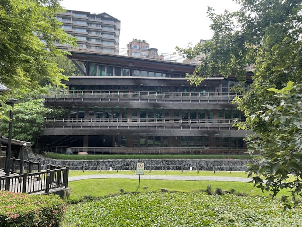 A modern multi-story building surrounded by greenery. The structure features horizontal lines and large glass windows, blending with the natural environment. There are trees and bushes in the foreground and background, providing a serene and natural setting.
