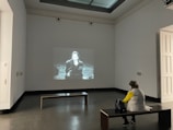 A person is sitting on a bench in a minimally decorated gallery room, facing a black-and-white movie projected onto the wall. The person appears to be watching the film intently. The room has high ceilings and some natural light coming in from the top.