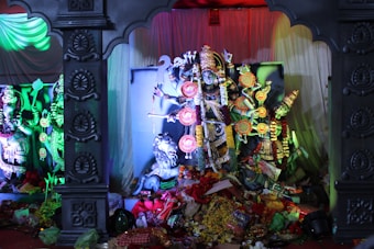In the center, there is a decorated statue of a deity with multiple arms and ornate attire, including colorful garlands and accessories. On each side, additional statues of deities are present. The backdrop is a combination of white fabric drapery with colorful lighting that creates a mystical ambiance. The floor is cluttered with various offerings such as flowers, fabrics, and other items.
