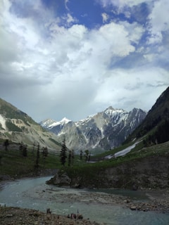A picturesque landscape features tall, snow-capped mountains under a partly cloudy sky. A river flows through the lush green valley, with scattered trees and rocks along its banks. A small group of people stand by the riverside, adding a sense of scale to the serene scene.