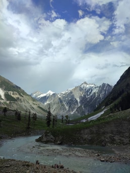 A picturesque landscape features tall, snow-capped mountains under a partly cloudy sky. A river flows through the lush green valley, with scattered trees and rocks along its banks. A small group of people stand by the riverside, adding a sense of scale to the serene scene.
