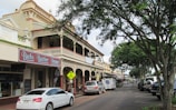 A bustling street lined with historic buildings featuring intricate architectural details and balconies. The scene captures multiple parked cars along the curb, a mix of modern and classic cars, with trees lining the opposite side of the street providing shade. A business sign for 'Bucks Butcher Shoppe' is prominently displayed on one building.