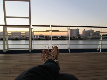 A relaxed person is lying on a deck chair, their feet up, wearing sandals. The view ahead shows an industrial area across a body of calm water, with large storage tanks and structures silhouetted against a serene sunset sky.