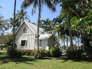 A charming small church with a light-colored wooden exterior is surrounded by lush greenery and tall palm trees. The church features a steep, metal roof and a cross at the top. The windows are elegantly shaped, adding to the beautiful, tranquil setting.