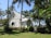 A charming small church with a light-colored wooden exterior is surrounded by lush greenery and tall palm trees. The church features a steep, metal roof and a cross at the top. The windows are elegantly shaped, adding to the beautiful, tranquil setting.