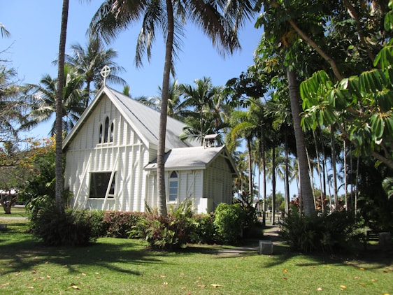 A charming small church with a light-colored wooden exterior is surrounded by lush greenery and tall palm trees. The church features a steep, metal roof and a cross at the top. The windows are elegantly shaped, adding to the beautiful, tranquil setting.
