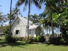 A charming small church with a light-colored wooden exterior is surrounded by lush greenery and tall palm trees. The church features a steep, metal roof and a cross at the top. The windows are elegantly shaped, adding to the beautiful, tranquil setting.