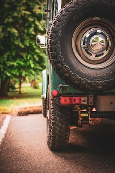 A close-up view of the rear end of an off-road vehicle, focusing on the large spare tire mounted on the back. The vehicle is parked on a paved path with lush green trees in the background, indicating a natural or park-like setting.