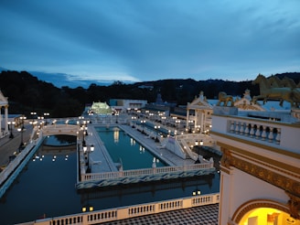 An expansive outdoor scene featuring a grand architectural complex with ornate structures adorned with horse sculptures. The scene is illuminated with numerous lamps, casting a soft glow over the decorative bridges, stairways, and water channels. The atmosphere is tranquil, framed by a deep blue evening sky.