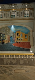 A detailed mural depicting a winding street with colorful buildings on both sides, leading to a large painting canvas set in an outdoor urban environment. The architecture is ornate, with decorative columns and arches framing the scene. The mural is illuminated by lights, enhancing its vibrant colors and intricate details.
