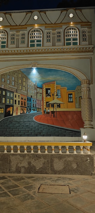 A detailed mural depicting a winding street with colorful buildings on both sides, leading to a large painting canvas set in an outdoor urban environment. The architecture is ornate, with decorative columns and arches framing the scene. The mural is illuminated by lights, enhancing its vibrant colors and intricate details.