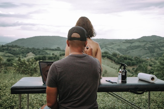 A man wearing a cap sits at a portable massage table outdoors, overlooking a lush, green hilly landscape. In front of him, a woman with short hair sits topless on the table, facing away towards the scenery. On the table are various items including a spray bottle labeled H2O, a roll of paper towels, and some small bottles and pens.