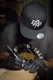 A person wearing a black cap and gloves is focused on tattooing someone’s skin. They have a concentrated expression and are holding a tattoo machine. The person is wearing a dark t-shirt with some graphic text and a bear illustration.
