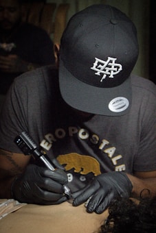 A person wearing a black cap and gloves is focused on tattooing someone’s skin. They have a concentrated expression and are holding a tattoo machine. The person is wearing a dark t-shirt with some graphic text and a bear illustration.
