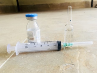 A syringe lies on a flat surface next to two small medicine vials, one with a blue cap. The syringe is marked with measurement increments, and the vials appear to contain liquid.