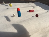 A collection of various pills including capsules and tablets is captured mid-air on a white cloth background. There are pills in different colors such as blue and red, yellow, white, and maroon. Shadows of the pills are cast on the surface, suggesting strong lighting from one direction.