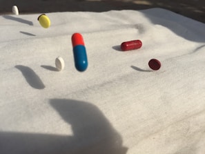A collection of various pills including capsules and tablets is captured mid-air on a white cloth background. There are pills in different colors such as blue and red, yellow, white, and maroon. Shadows of the pills are cast on the surface, suggesting strong lighting from one direction.