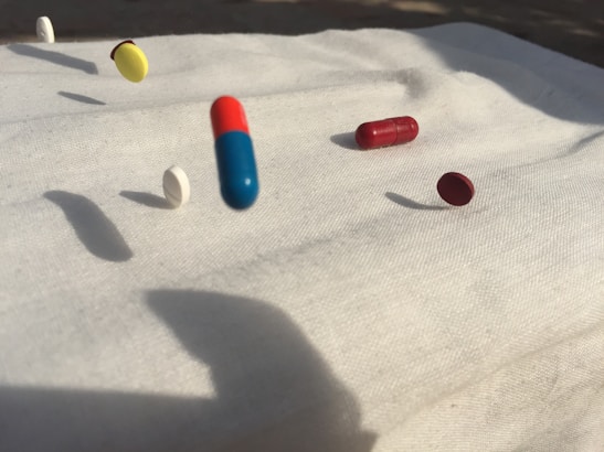 A collection of various pills including capsules and tablets is captured mid-air on a white cloth background. There are pills in different colors such as blue and red, yellow, white, and maroon. Shadows of the pills are cast on the surface, suggesting strong lighting from one direction.