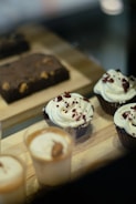 Cupcakes with creamy white frosting and red sprinkles are displayed alongside small cups with a caramel-colored layer and a chocolate brownie with visible nuts on a wooden tray.