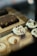 Cupcakes with creamy white frosting and red sprinkles are displayed alongside small cups with a caramel-colored layer and a chocolate brownie with visible nuts on a wooden tray.
