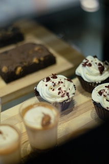 Cupcakes with creamy white frosting and red sprinkles are displayed alongside small cups with a caramel-colored layer and a chocolate brownie with visible nuts on a wooden tray.